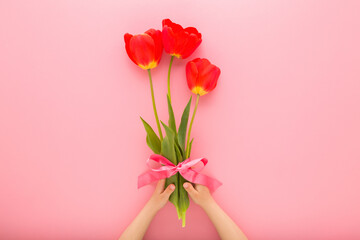 Little baby girl hands holding fresh beautiful red tulip flowers with green leaves and ribbon on...
