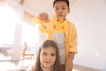 Cute children of different races play together. A small Asian boy holds a paper crown over the head...