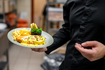chef holds a plate of a delicious meal, featuring a creamy dish garnished with herbs and spices, accompanied by golden fries and fresh vegetables