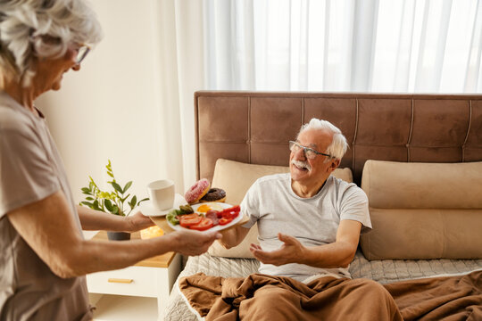 Romantic senior woman is bringing breakfast in bed to her husband while standing in a bedroom.