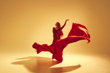 Artistic and deep performance Woman, female dancer in flowing red dress dancing against golden color background. Concept of art, classical dance, beauty and fashion, aesthetics