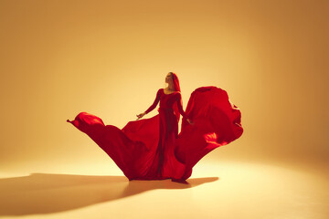 Artistic woman in elegant red flowing dress making creative performance, dancing against golden...