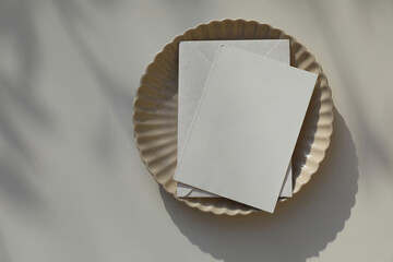 Blank greeting card, invitation mockup, envelope in sunlight. Scalloped ceramic plate on beige...