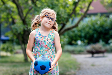Little preschool girl with eyeglasses playing with ball outdoors. Happy smiling child catching and throwing, laughing and making sports. Active leisure with children and kids. Summer day on backyard