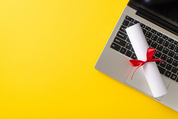 An image of a rolled-up diploma tied with a red ribbon on a laptop keyboard, set against a yellow...