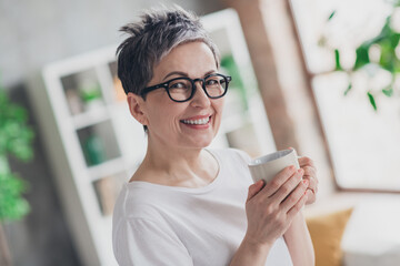 Photo of lovely mature lady drink coffee dressed white garment modern living room rest enjoy...