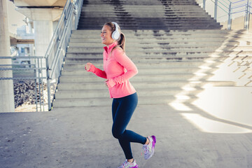 Exercise, music and a sports woman running in the city for health or cardio preparation of a marathon. Fitness, wellness or training and a young runner or athlete listening to audio with headphones.