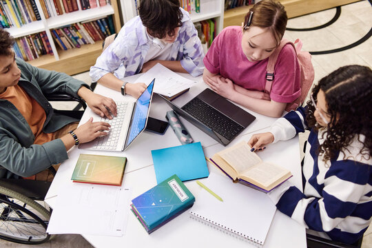 High angle view of diverse group of students sitting at table in school library together and doing research with textbooks and computers copy space