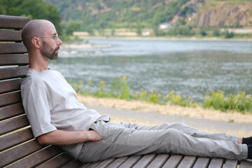 young man 30 years old sites comfortably on wooden park bench on banks of river, lake, resting on...