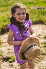 A little girl in a purple dress, with wavy hair and two ponytails in a straw hat walks in a field...