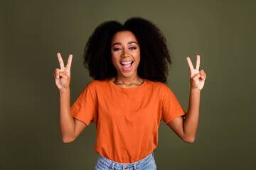 Portrait of overjoyed cheerful girl with wavy hairstyle wear orange t-shirt showing v-sign symbol isolated on dark khaki background