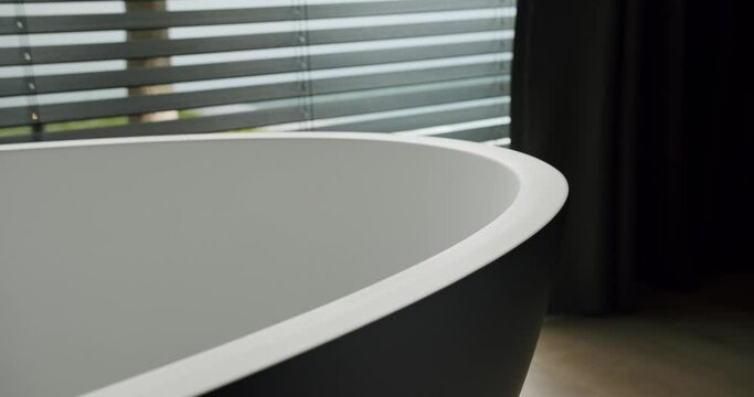 Closeup of bath tub near large window with blinds and curtains in luxury apartment with modern interior. Minimalist design in bathroom. Bath tub in luxury apartment with modern interior