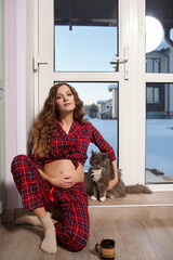 A pregnant woman in the living room with cup of tea sits on the threshold of a glass door and plays...