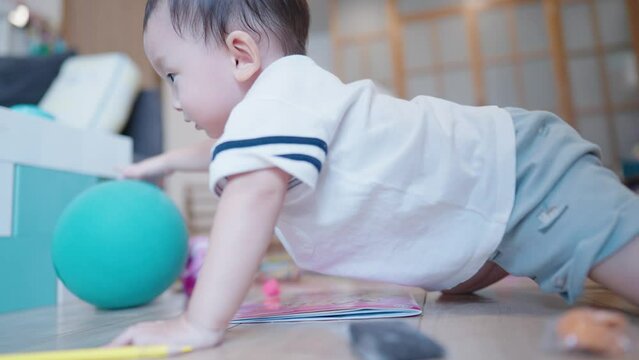 Happy asian baby crawls in the room playing with blue rubber air ball next to cozy sofa. Boy Toddler playing in the house and learns how to crawl, child development skills, learning age preschooler 