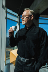 A gray-haired man in a black shirt and coffee in his hands stand by the window in the office.