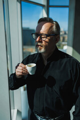 A gray-haired man in a black shirt and coffee in his hands stand by the window in the office.