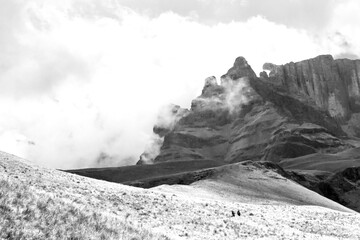 Distant hikers walking towards the ominous, mist covered peaks of the Drakensberg Mountains of...