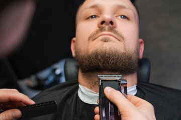 Master cuts client beard with automatic trimmer in barbershop closeup. Hairstylist does haircut with modern equipment in grooming salon