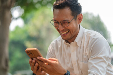 Smiling adult businessman sitting in the park and using mobile phone on on sunny day.