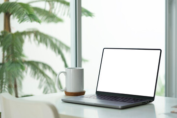 Laptop computer with blank screen, notepad and cup of coffee on white working desk near window.