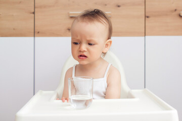 Unhappy baby sitting in a baby chair in the kitchen. Child drinks water. Feeding and childhood