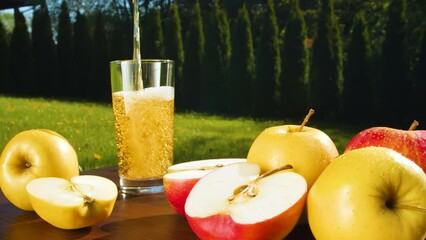 Close-up of apple juice being poured into a glass surrounded by red and yellow apples. Circular filming. Outdoors.