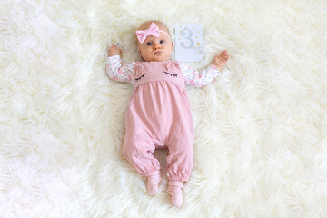 A baby girl, wearing a pink onesie and a bow on her head, lies on a soft, white blanket with a card...