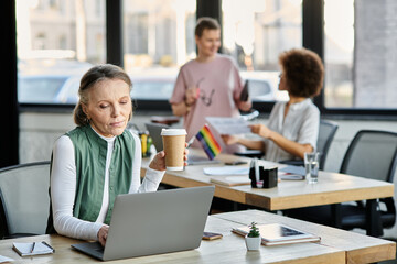 Beautiful mature woman works on a laptop at a table, with her diverse colleagues on backdrop.