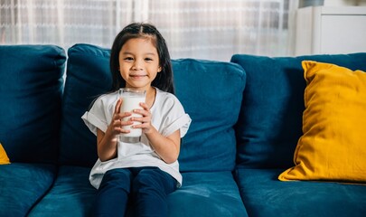 An adorable Asian child enjoys a glass of milk on the comfy sofa at home radiating happiness and...