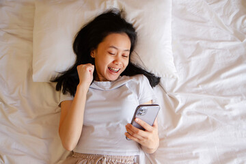 Top view of happy asian young woman awaken in white cozy bed using modern smartphone gadget, happy...