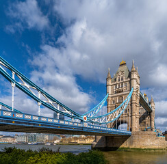 Obraz premium View from the edge of the River Thames of London's Tower Bridge illuminated by the morning sun under a blue sky with white clouds and ferry traffic on the river and docks in the background. United Kin