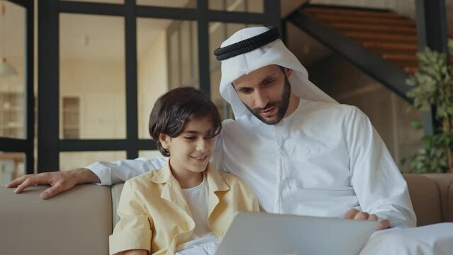 Father and son studying together on the couch. Man from emirati wearing kandura traditional dress teaching to his son on the laptop.