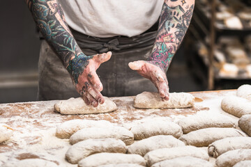 A baker with tattoos forms raw dough buns in the bakery