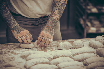 A baker with tattoos forms raw dough buns in the bakery