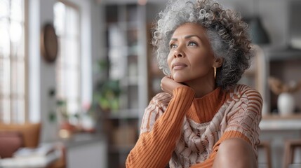 A thoughtful senior biracial woman indoors, gazing upwards with a hand on her chin, in a cozy home setting.