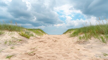 A tranquil sandy hill pathway leading to the ocean, framed by green grass under a cloudy sky.