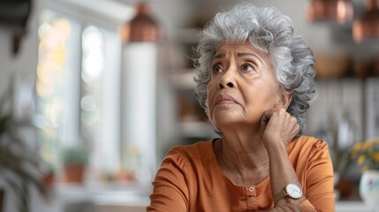A senior biracial woman appears contemplative, sitting inside a home with a thoughtful expression.