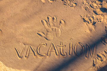 At sunset the inscription on the white sand vacation with a handprint on the sea coast. Beautiful white sand on the beach close-up in the rays of the setting sun with the inscription vacation.