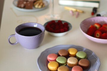Plate of pastel macarons, cookies and chocolate, cup of tea of coffee, glass of bubble water, various berries, books and accessories on the table. Selective focus, pastel colors.