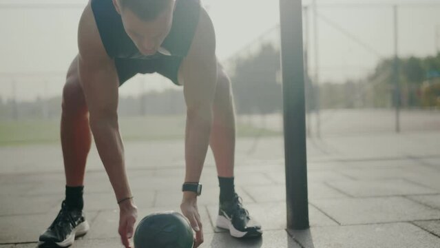 Determined male athlete in intense outdoor fitness session stretches with a medicine ball, showcasing strength and flexibility. Focused Young Man in Outdoor Workout Stretching with Medicine Ball