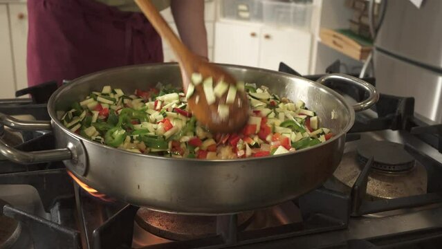 man hand stirring sofrito in large frying pan with sofrito of onion, pepper and garlic. Cooking with gas and dressed in maroon apron and green t-shirt.