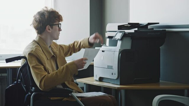 Tilt up shot of young male office worker riding wheelchair to printer and copying business documents on it during workday