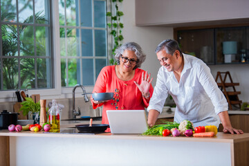 Indian asian senior old age cheerful couple using tablet computer or laptop in the kitchen while cooking