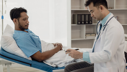 Obraz premium A young man lies on a bed in a hospital ward talking with her young Indian doctor. Doctor checking patient's health doctor treating patient, hospital admission of patient