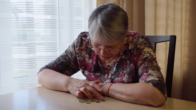 An elderly woman sits alone by the window and longingly counts small money, coins in euros. Poverty of pensioners and the elderly.