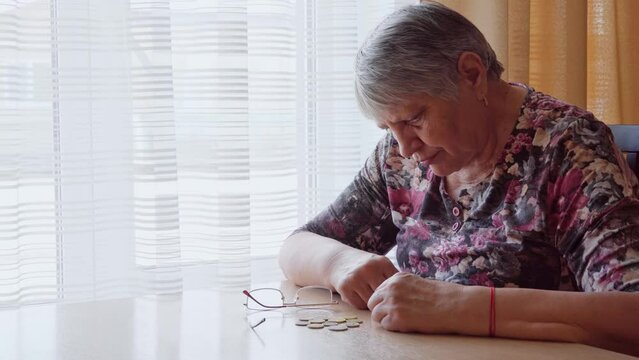 An elderly woman sits alone by the window and longingly counts small money, coins in euros. Poverty of pensioners and the elderly.