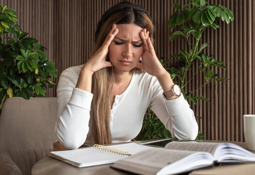 Focused yet stressed adult woman studying at home, showing signs of ADHD, dyslexia, and learning difficulties, sitting at a desk surrounded by books and lush greenery 