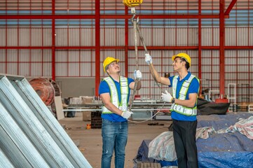 Two workers wearing hard hats and safety vests use a chain hoist to lift a heavy metal beam in a...