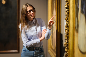Thoughtful young Caucasian woman wearing glasses and looking at exhibition. Concept of Museum Day.