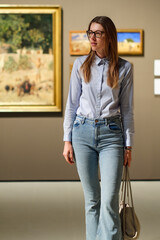 Thoughtful young Caucasian woman wearing glasses and looking at exhibition. Concept of Museum Day.